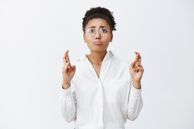 La femme supplie Dieu que le rêve devienne réalité. Portrait de femme bouleversée de mendicité nerveuse dans des verres et une chemise blanche, levant les lèvres, pincer les lèvres et croiser les doigts pour prier, croyant aux miracles en espérant