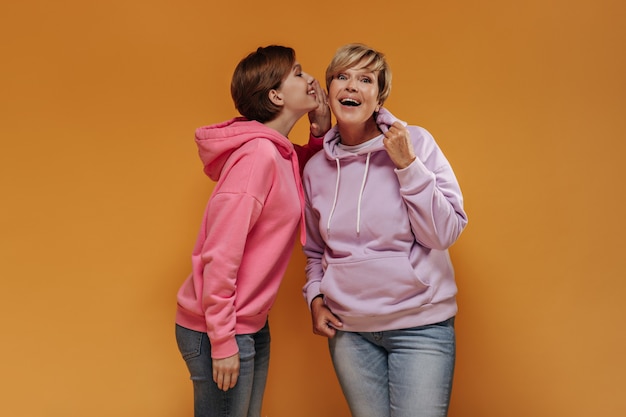 Femme surprise aux cheveux blonds en sweat-shirt lilas et jeans écoutant le secret de la petite-fille sur fond orange.