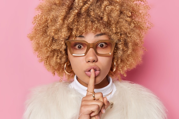 Photo gratuite une femme surprise aux cheveux bouclés et touffus fait un geste de silence dit que le secret porte des lunettes et un manteau de fourrure blanc isolé sur fond rose. chut ne répandez pas de rumeurs s'il vous plait. notion de langage corporel