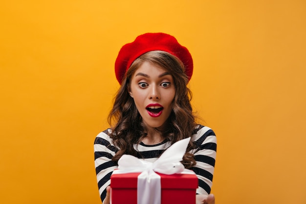 Femme surprise en béret rouge, obtenant une boîte-cadeau. Curly jeune fille avec rouge à lèvres brillant en chapeau posant sur fond isolé.