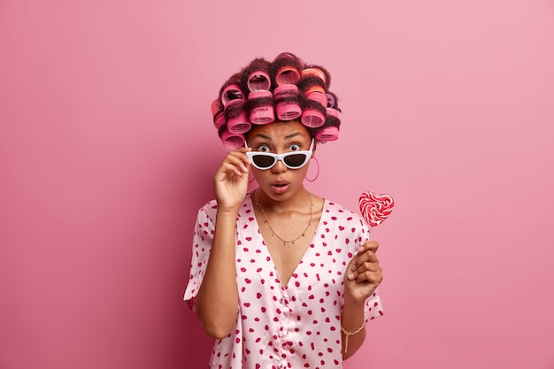 Photo gratuite femme surprise avec des bigoudis dans les cheveux, se prépare pour la journée des femmes, veut avoir un look brillant, porte un peignoir et des lunettes de soleil, tient une délicieuse sucette appétissante, isolée sur un mur rose