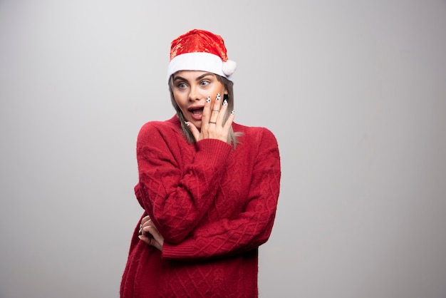 Femme surprise en bonnet de Noel debout sur fond gris.