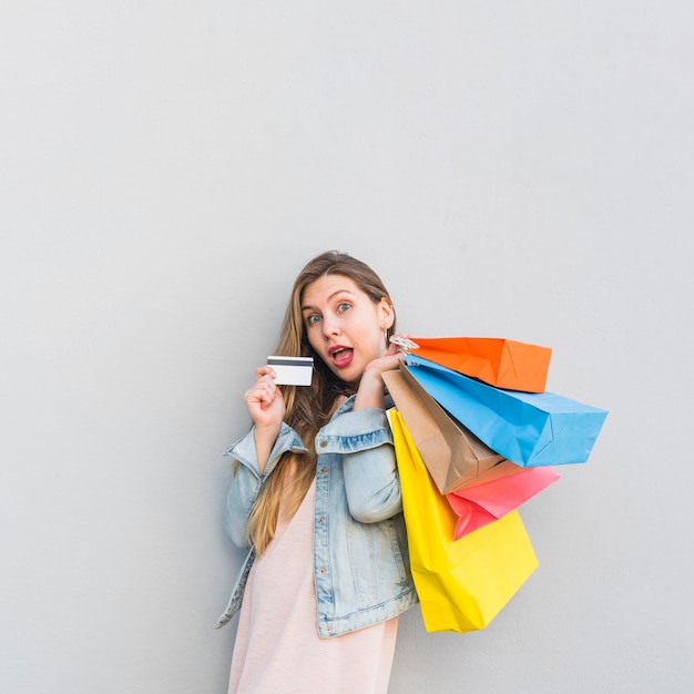 Femme surprise debout avec des sacs à provisions et une carte de crédit au mur