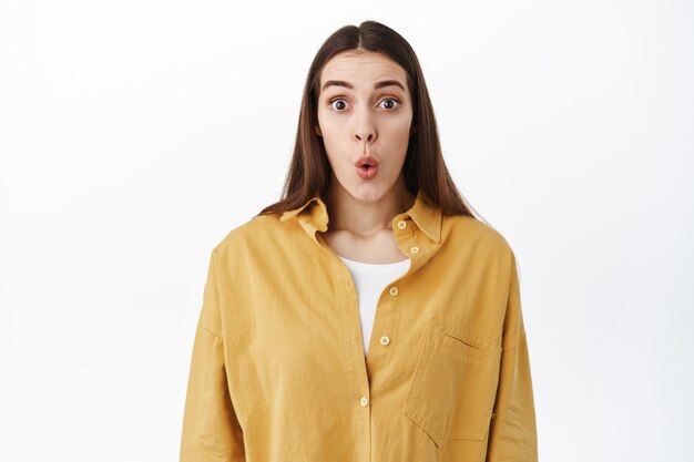 Une femme surprise et excitée lève les sourcils et plie les lèvres impressionnée, regarde quelque chose de génial, regarde une offre promotionnelle incroyable, a l'air fascinée, debout sur un mur blanc