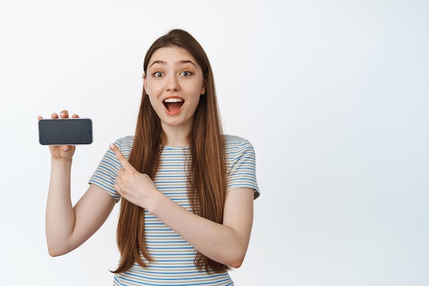 Femme surprise et heureuse pointant le doigt sur l'écran horizontal du smartphone, souriant étonné, montrant la publicité de l'application, le site Web sur le téléphone mobile, fond blanc.