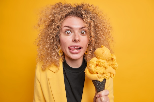 Une femme surprise joyeuse avec des cheveux bouclés et touffus lèche les lèvres qui sort la langue tient de grosses glaces délicieuses tentant de manger des poses de desserts d'été contre un mur jaune vif. Mmm comme c'est délicieux.