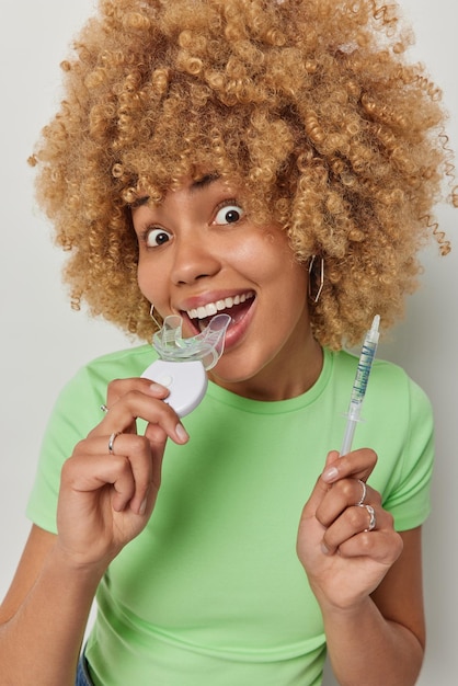 Une femme surprise positive aux cheveux blonds et bouclés tient un extenseur de bouche en plastique et une seringue avec du gel pour l'appliquer porte un t-shirt vert décontracté pose à l'intérieur sur fond blanc Personnes et soins bucco-dentaires