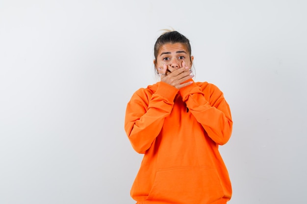 Femme en sweat à capuche orange gardant les mains sur la bouche et semblant choquée