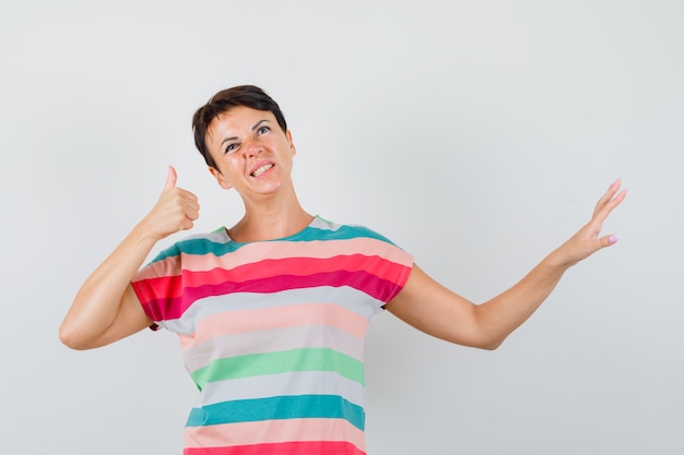 Femme en t-shirt rayé montrant le geste d'arrêt avec le pouce vers le haut et à la vue de face, hésitante.