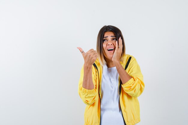 Femme en t-shirt, veste montrant le pouce vers le haut, gardant la main sur la joue et regardant excité, vue de face.