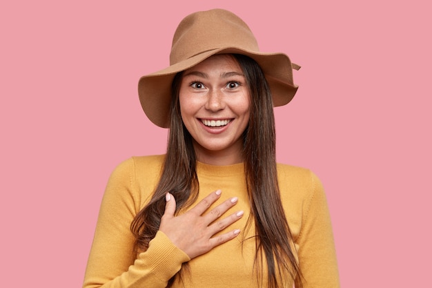 Femme de taches de rousseur surpris émotionnel avec expression positive garde la main sur la poitrine, sourit largement à la caméra