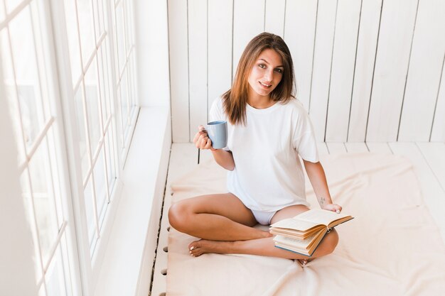 Femme avec une tasse et un livre en regardant la caméra