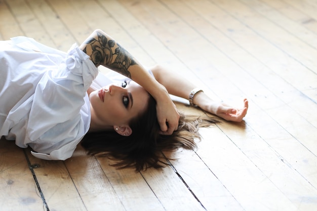 Femme tatouée dans une chemise blanche allongée sur le plancher en bois