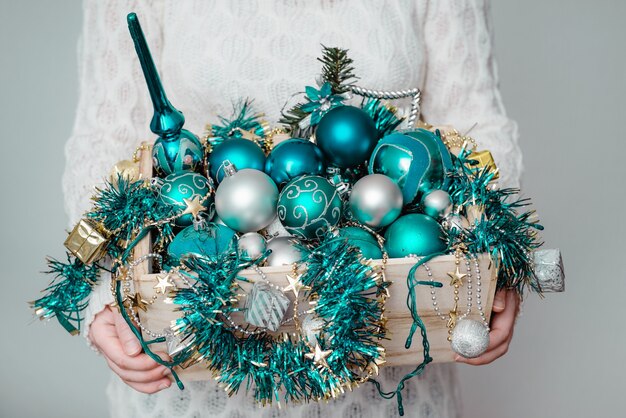 Femme tenant une boîte de guirlandes et ornements de Noël bleu sur mur gris