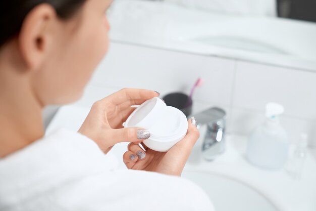 Femme tenant la crème pour le visage ou le corps dans la salle de bain