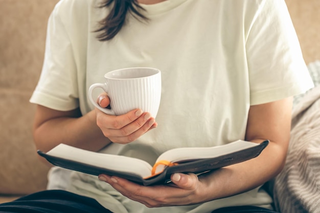 Photo gratuite une femme tenant une tasse de thé et lisant un livre sur le canapé à la maison de près