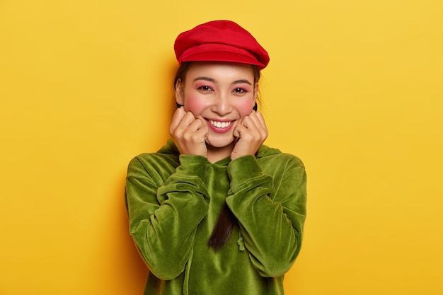 Femme tendre charismatique d'apparence orientale, touche les joues, profite de la meilleure journée, porte une casquette rouge vif et un sweat-shirt vert en velours