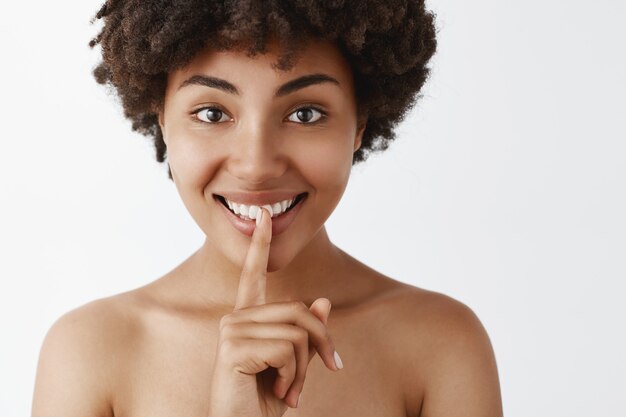 Femme tendre et féminine à la peau foncée avec une peau propre et douce, souriant avec une expression intrigante, montrant un signe de chut avec l'index sur la bouche