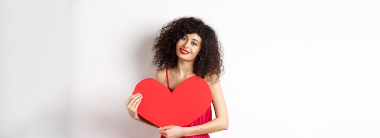 Photo gratuit femme tendre romantique avec des cheveux bouclés serrant un grand coeur rouge et un regard souriant avec amour debout sur fond blanc