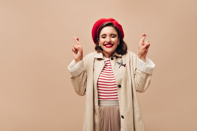 Femme en tenue d'automne élégante croise les doigts sur fond beige. Jeune fille souriante en béret rouge et en manteau à la mode posant.