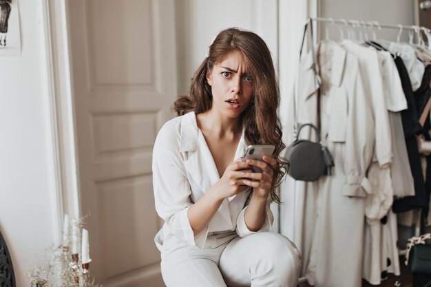 Une femme en tenue blanche tient un téléphone et a l'air choquée