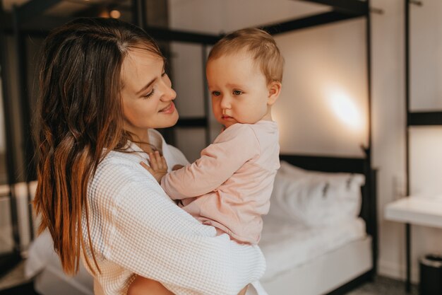 Femme en tenue de maison blanche regarde l'enfant et la tient dans les bras. Fille touche maman sur fond de lit.