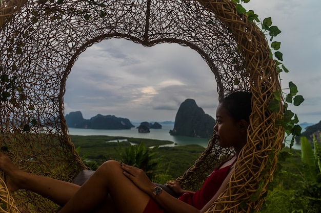 Photo gratuite femme avec une tenue rouge assis sur une balançoire en osier tissé près de la mer sous les nuages d'orage