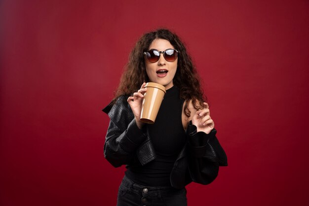 Femme en tenue toute noire posant avec une tasse et des lunettes.