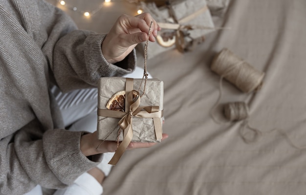 Une femme tient une boîte-cadeau de Noël décorée dans un style artisanal, décorée de fleurs séchées et d'une orange sèche, enveloppée dans du papier kraft.