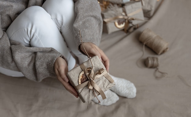 Une femme tient une boîte-cadeau de Noël décorée dans un style artisanal, décorée de fleurs séchées et d'une orange sèche, enveloppée dans du papier kraft.