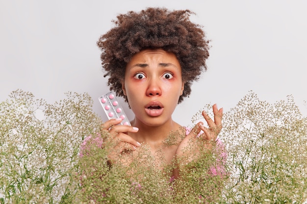 Photo gratuite une femme tient des pilules pour soigner l'allergie entourée d'un bouquet de fleurs sauvages a les yeux gonflés rouges se dresse sur blanc