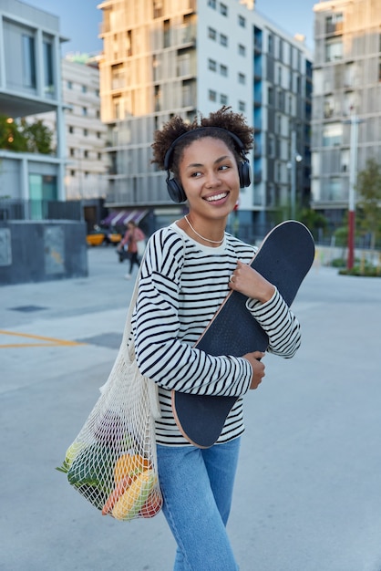 Photo gratuite une femme tient une planche à roulettes dans un sac en filet avec des légumes écoute de la musique en se promenant dans les rues de la ville vêtue d'un pull rayé et d'un jean profite de ses loisirs