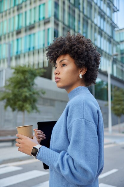 une femme tient une tablette numérique et une tasse de café en papier marche sur la route contre les bâtiments modernes de la ville en verre revient de l'université après des conférences habillées avec désinvolture