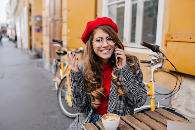 Femme timide avec une coiffure frisée posant dans un café en plein air avec sourire en septembre