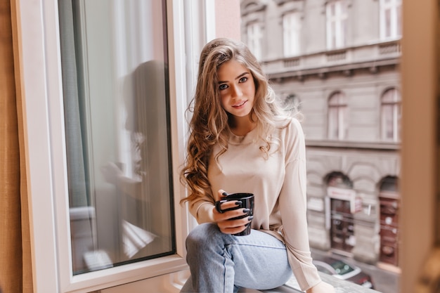 Femme timide en jeans assis sur le rebord de la fenêtre et posant avec intérêt