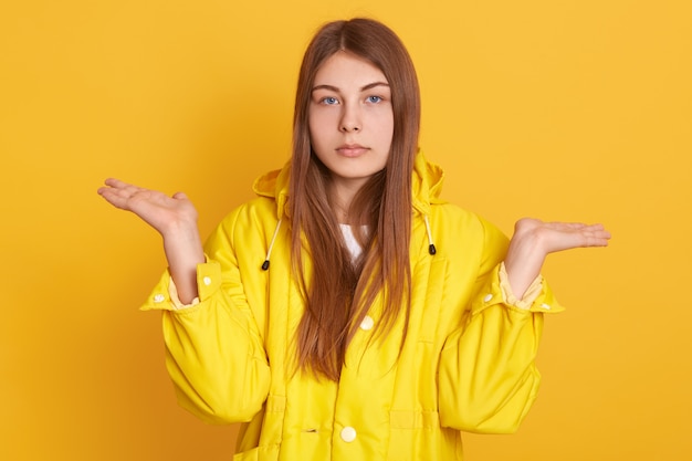 Femme triste caucasienne, écartant les mains, montrant un geste impuissant, une dame portant une veste, posant contre le mur jaune, a un air sérieux, avec une expression bouleversée.