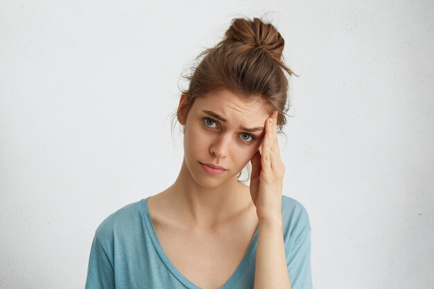 Femme triste déprimante avec noeud de cheveux, yeux bleus épuisés touchant sa tête ayant une expression mécontente d'être fatigué