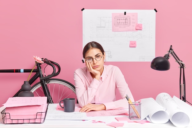 Une femme triste et fatiguée porte des lunettes sur le bureau et travaille toute la journée sur des plans impliqués dans le processus d'apprentissage