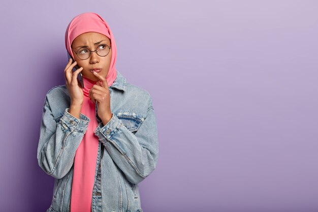 Femme triste à la peau sombre insatisfaite enveloppée dans un hijab rose, porte une veste en jean, des lunettes rondes