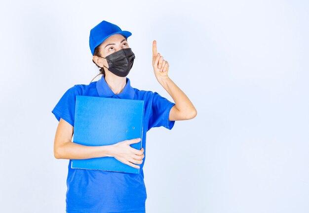 Femme en uniforme bleu et masque noir tenant un dossier bleu et pointant quelqu'un de côté.