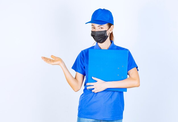 Femme en uniforme bleu et masque noir tenant un dossier bleu et pointant quelqu'un de côté.