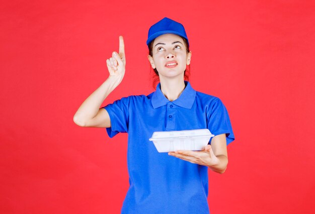 Femme en uniforme bleu tenant une boîte à emporter en plastique blanc pour la livraison et montrant quelque chose.