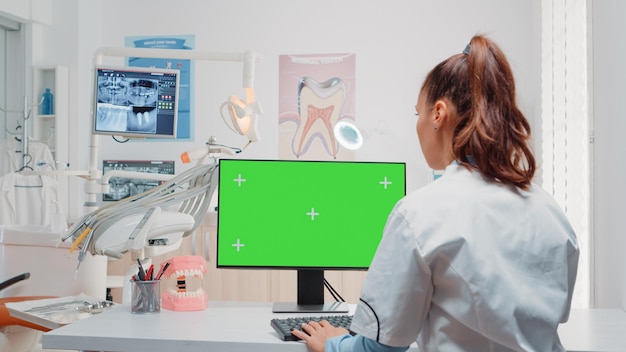 Femme utilisant un clavier et un ordinateur avec écran vert dans un cabinet dentaire pour les soins bucco-dentaires. dentiste travaillant avec chroma key sur moniteur avec modèle de maquette isolé pour la dentition et la dentisterie
