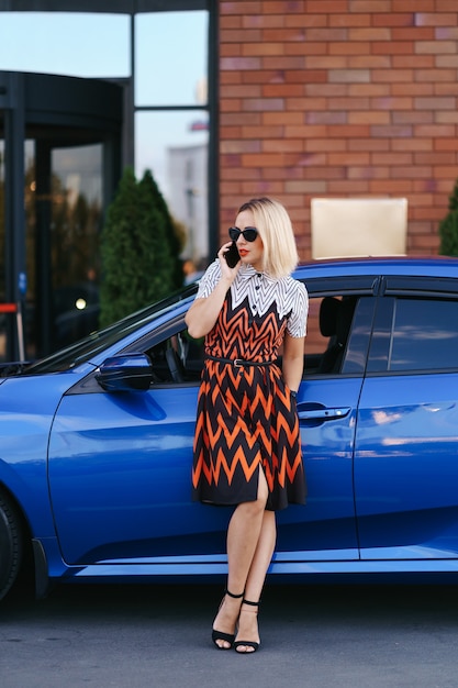 Femme utilisant un téléphone portable, une communication ou une application en ligne, debout près de la voiture sur la rue de la ville ou un parking, à l'extérieur. Autopartage, service de location