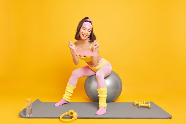 une femme utilise des accessoires de sport fait de l'exercice entraîne les muscles des bras avec une bande de résistance est assise sur un ballon de fitness vêtu de vêtements de sport