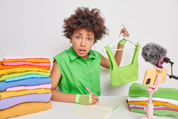 une femme vend des vêtements en ligne tient un haut recadré sur des cintres écrit des informations dans le bloc-notes est assise à table avec des piles de linge plié isolé sur blanc