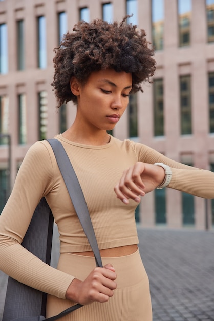 femme vérifie l'heure sur smartwatch après entraînement cardio va commencer l'exercice de pilates vêtu d'un survêtement beige se promène en milieu urbain