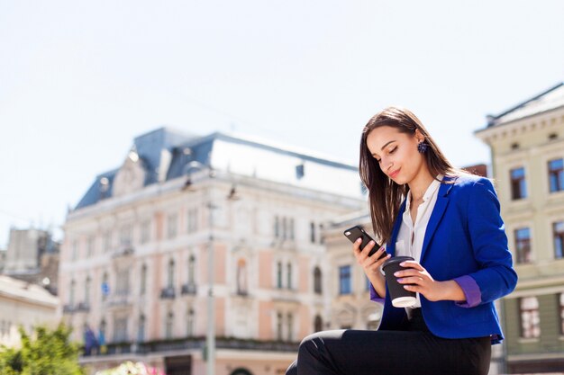 Femme vérifie son téléphone assis avec une tasse de café dans la rue