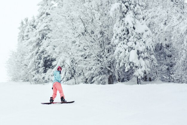 Femme en veste de ski bleu et pantalon rose se dresse sur le snowboard quelque part dans la forêt d&#39;hiver