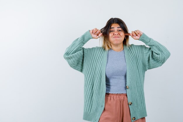 Femme en vêtements décontractés, brancher les oreilles avec les doigts, souffler les joues et regarder perplexe, vue de face.